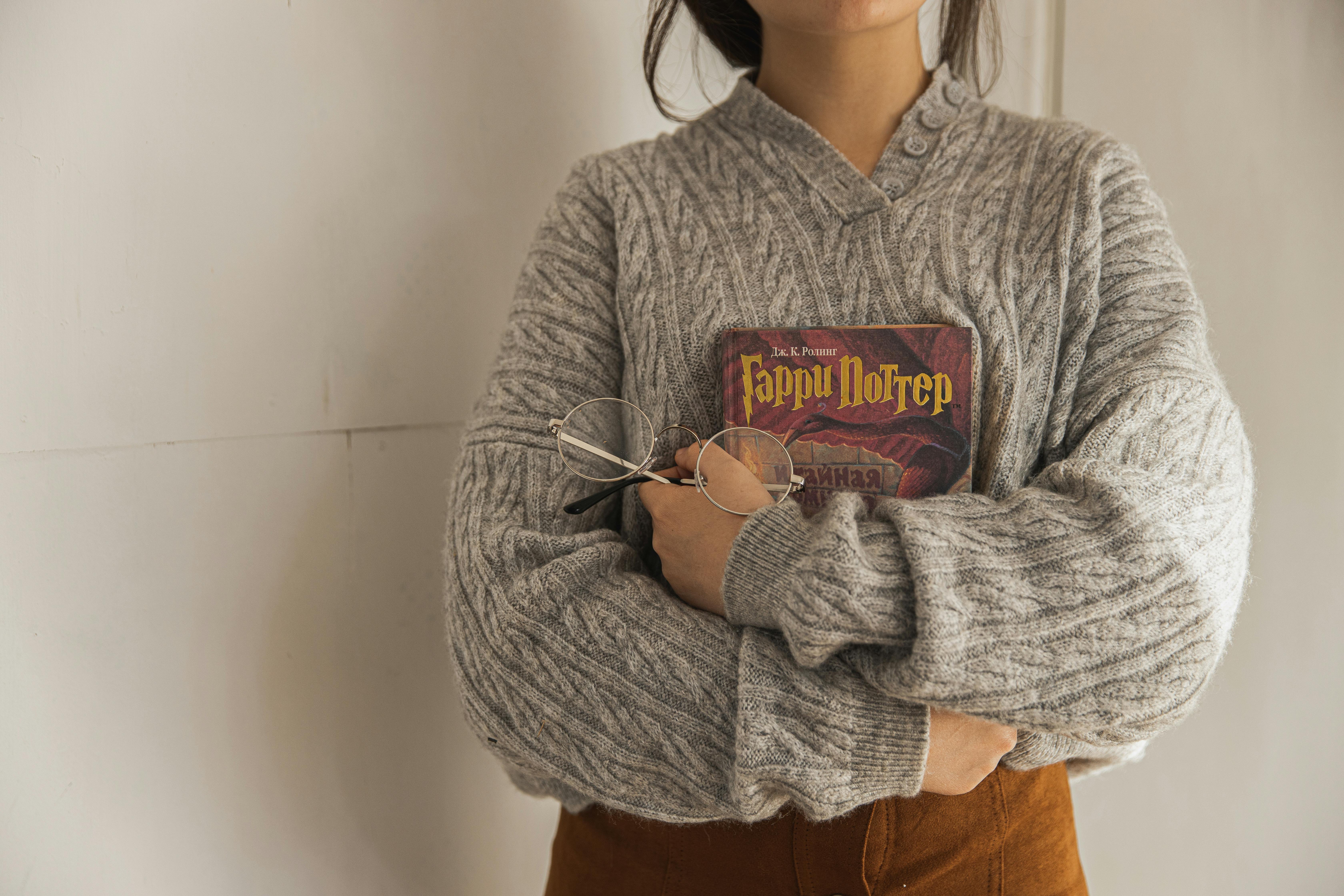 Woman in knit sweater holding a book and eyeglasses indoors on a fall day.