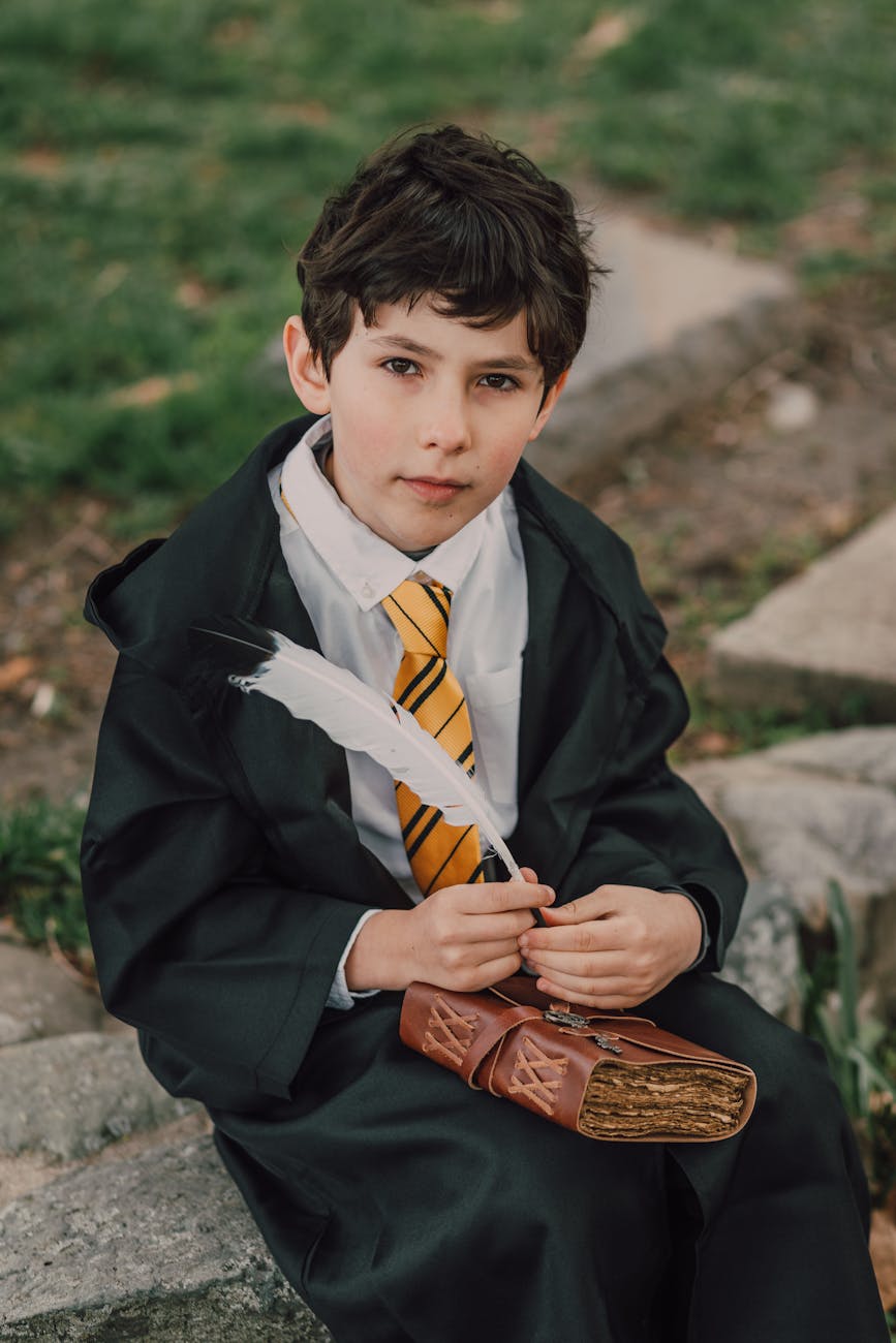 a boy in black robe holding a spell book and a quill